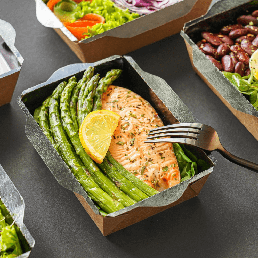 Grilled salmon with asparagus and a lemon slice in a black meal prep container, with a fork on the right side. - Home Instead