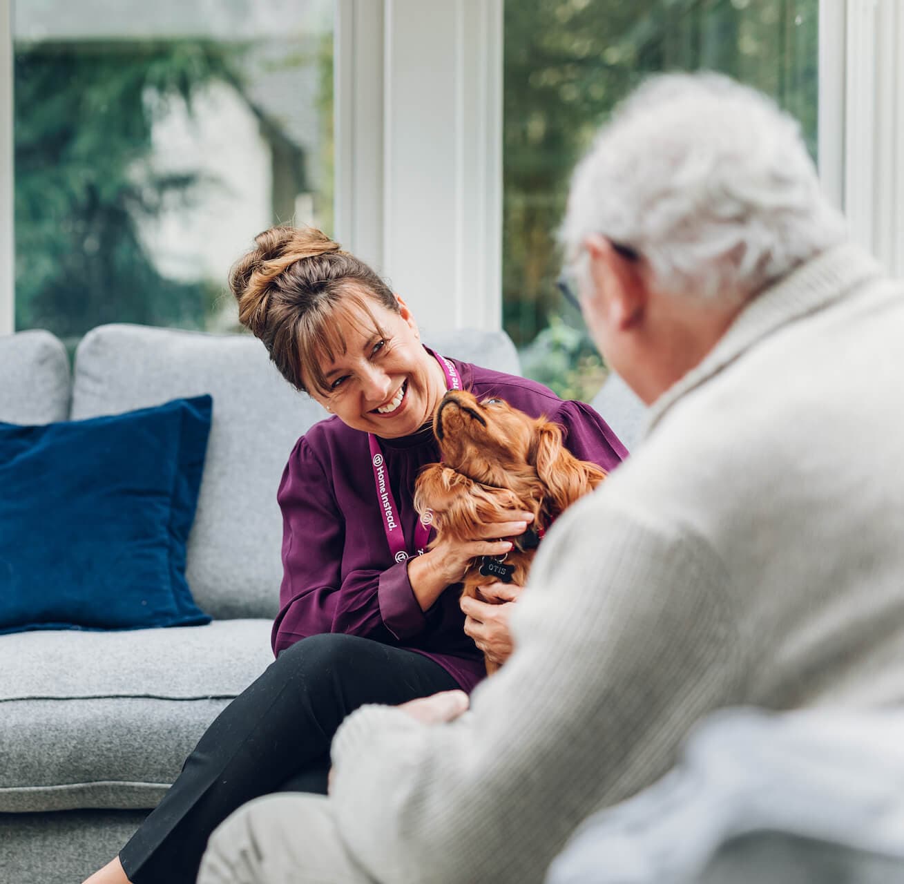 Woman smiling and holding a dog while talking to an elderly man seated on a couch in a bright living room. - Home Instead