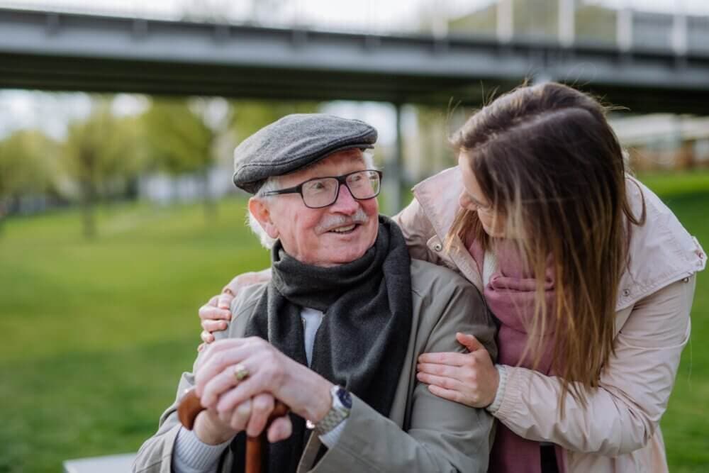 An elderly man with a cane and a young woman share a joyful moment outdoors, with a bridge and greenery in the background. - Home Instead Bournemouth & Christchurch
