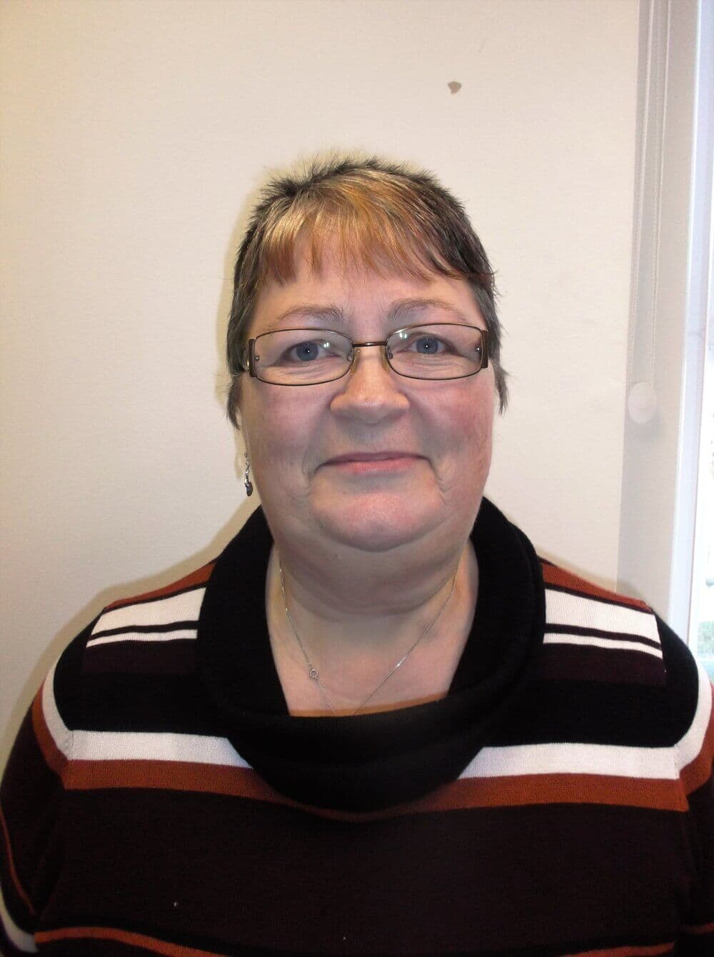 A woman with short hair and glasses, wearing a striped top, smiles while standing in front of a white wall. - Home Instead