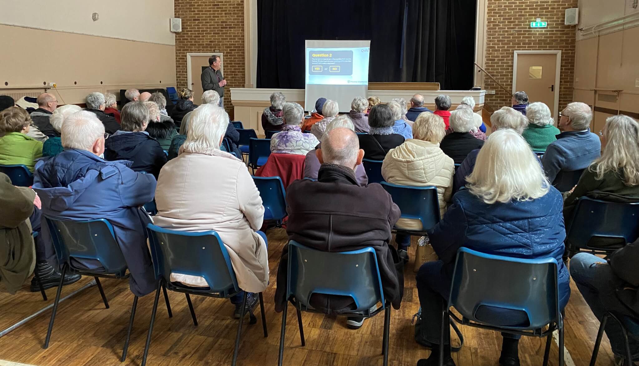 A group of people seated in a hall, watching a presentation on a screen at the front of the room. - Home Instead