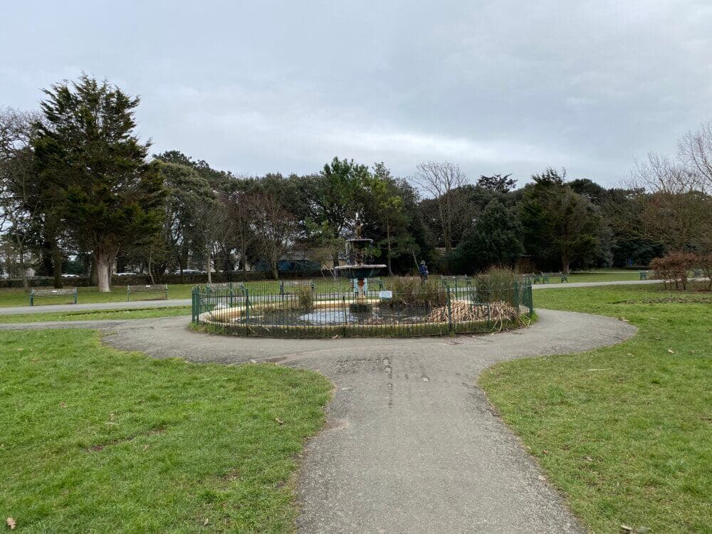 A small circular fountain surrounded by grass and trees in a park, with a paved path leading to it under a cloudy sky. - Home Instead