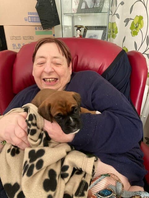 A joyful person sits in a red chair, holding a small brown puppy wrapped in a paw-print blanket. - Home Instead