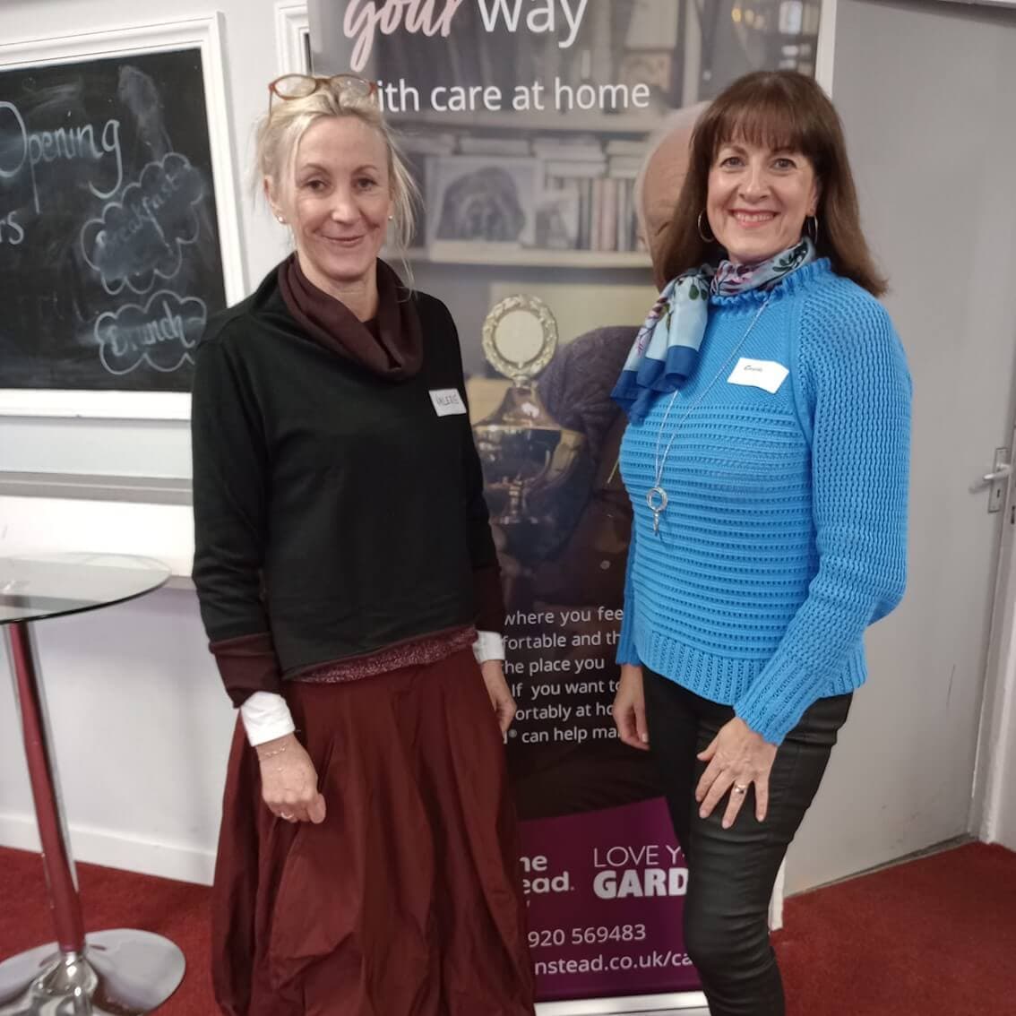 Two women smiling and standing in front of a care service banner in a room with red carpet and a whiteboard. - Home Instead