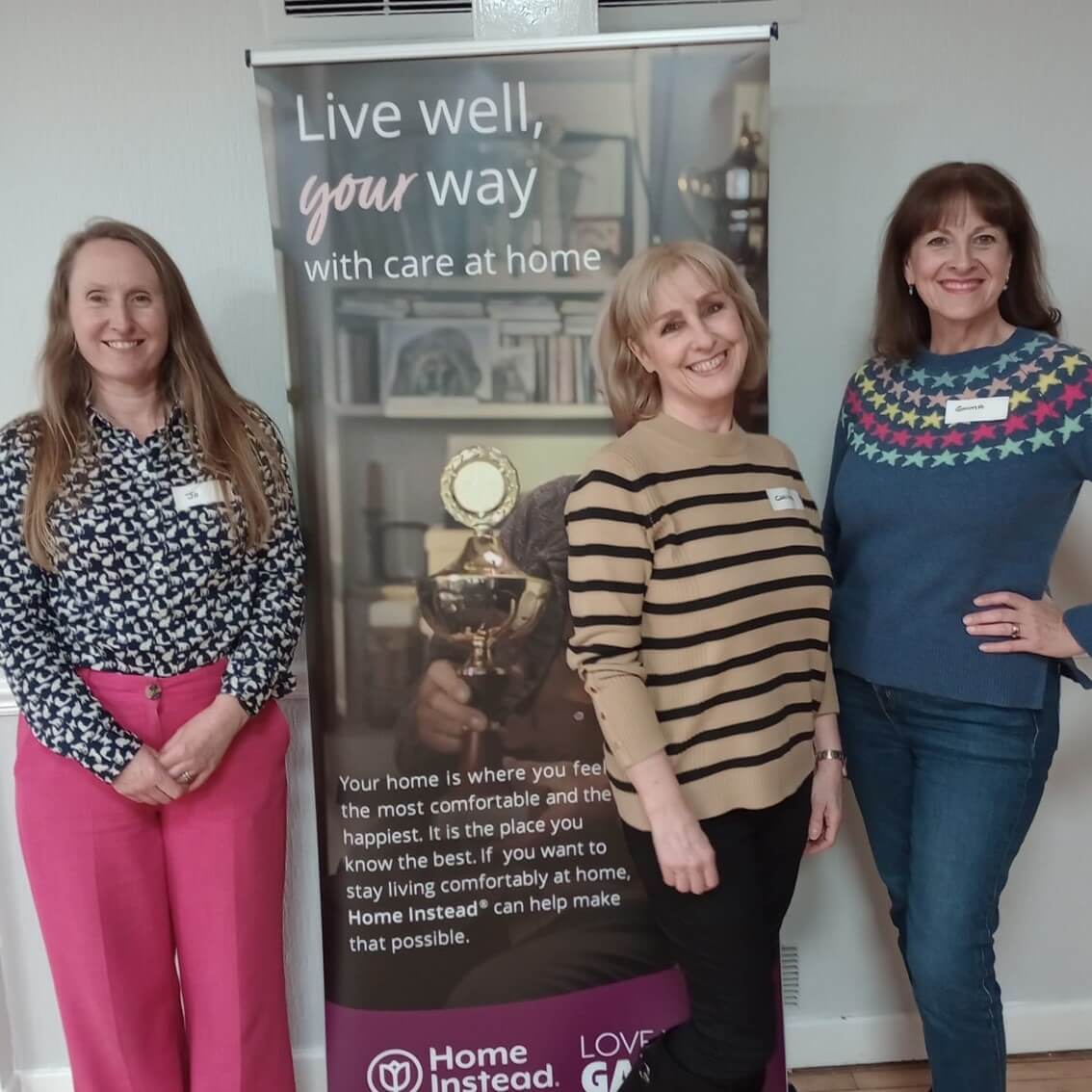 Three women standing in front of a banner that reads "Live well, your way with care at home," smiling at the camera. - Home Instead