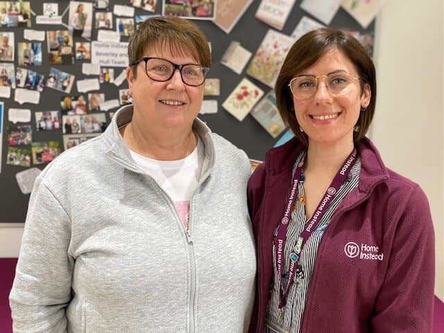 Two women are standing side by side, smiling. The woman on the right wears a Home Instead jacket. - Home Instead