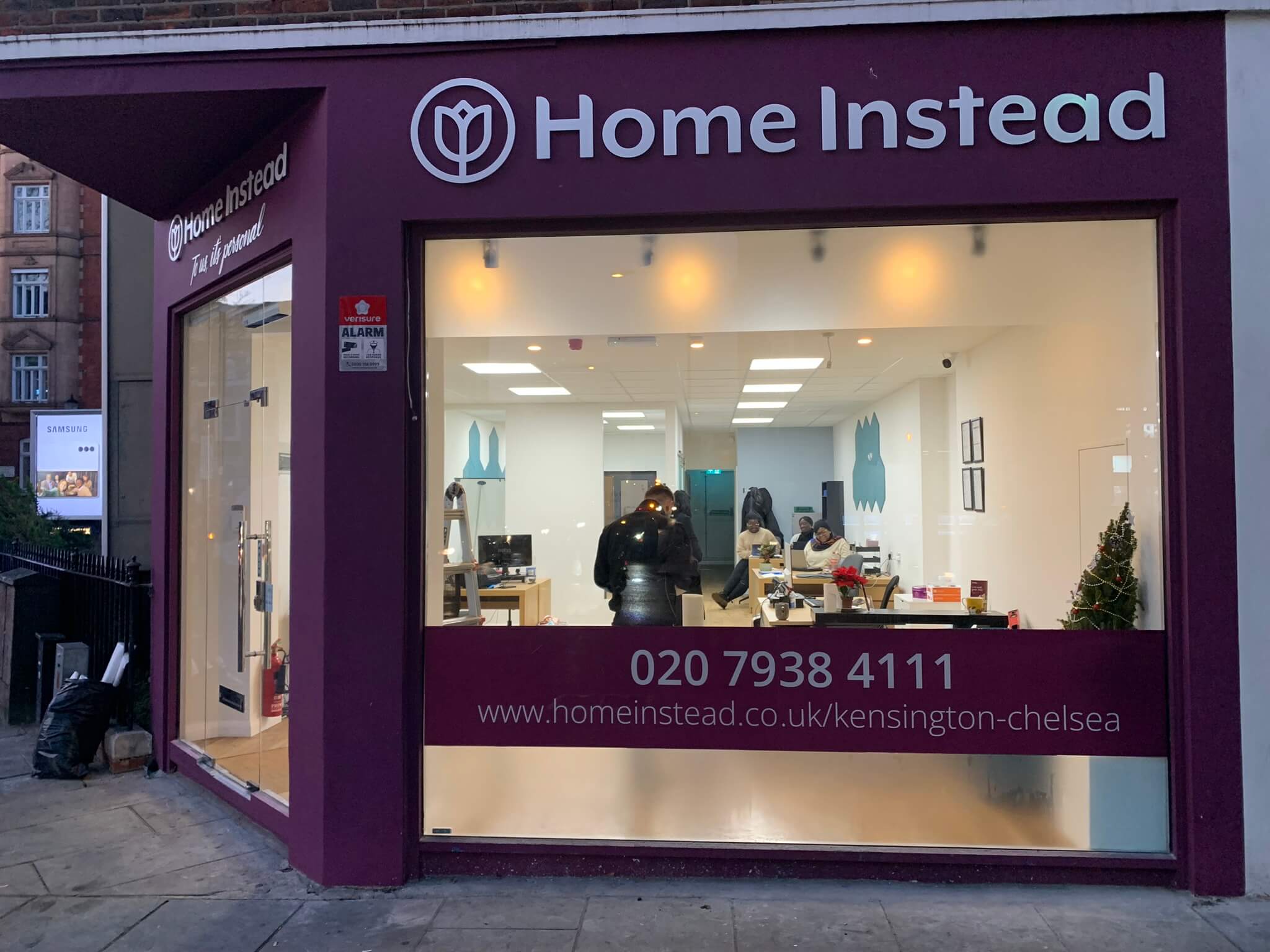 Exterior of Home Instead office with glass front, showing people inside working, and contact details on the window. - Home Instead