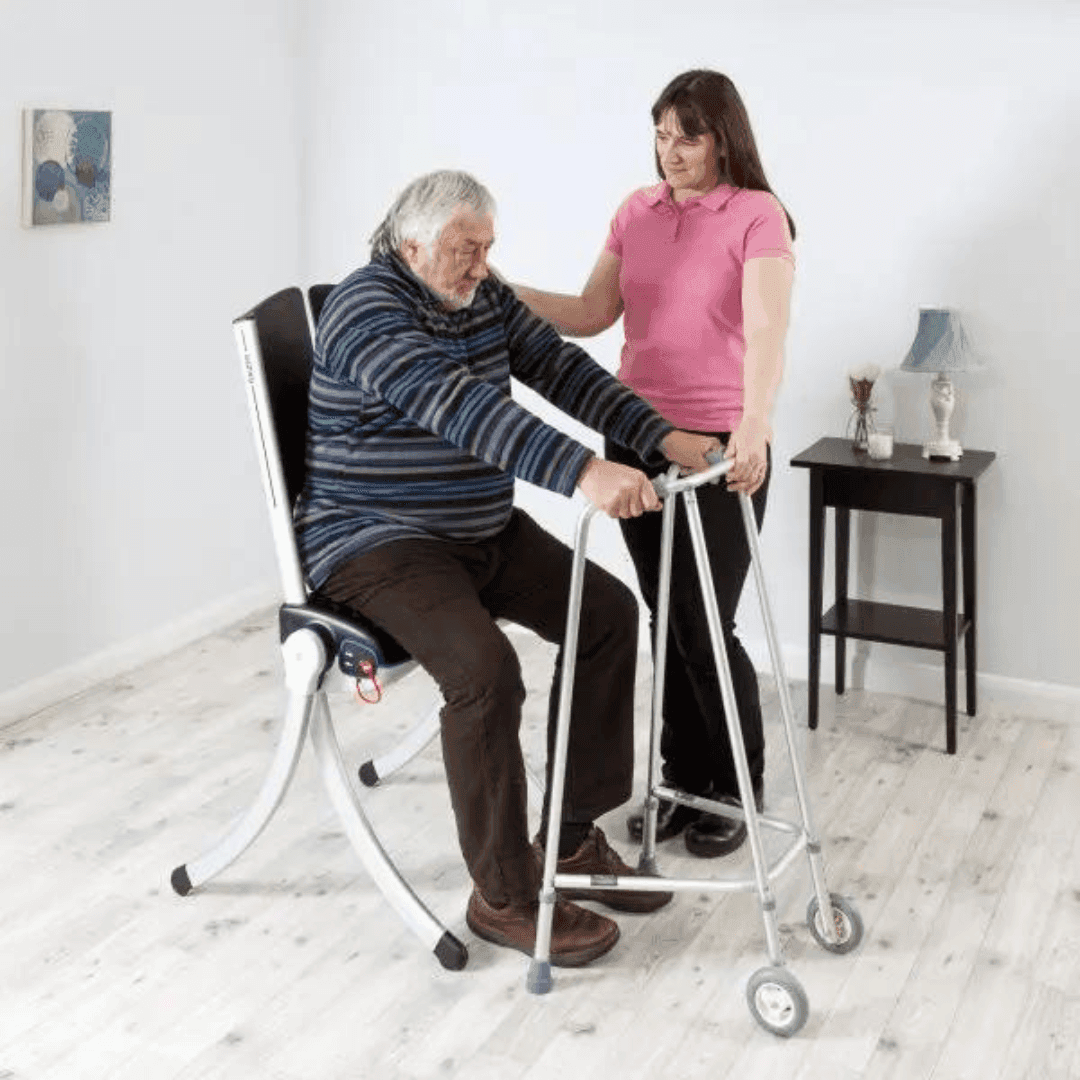 An elderly man sits on a lift chair holding a walker, assisted by a woman in a pink shirt in a bright room. - Home Instead