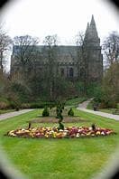 A church with tall steeples stands beyond a garden with vibrant flower beds and green lawns on a cloudy day. - Home Instead
