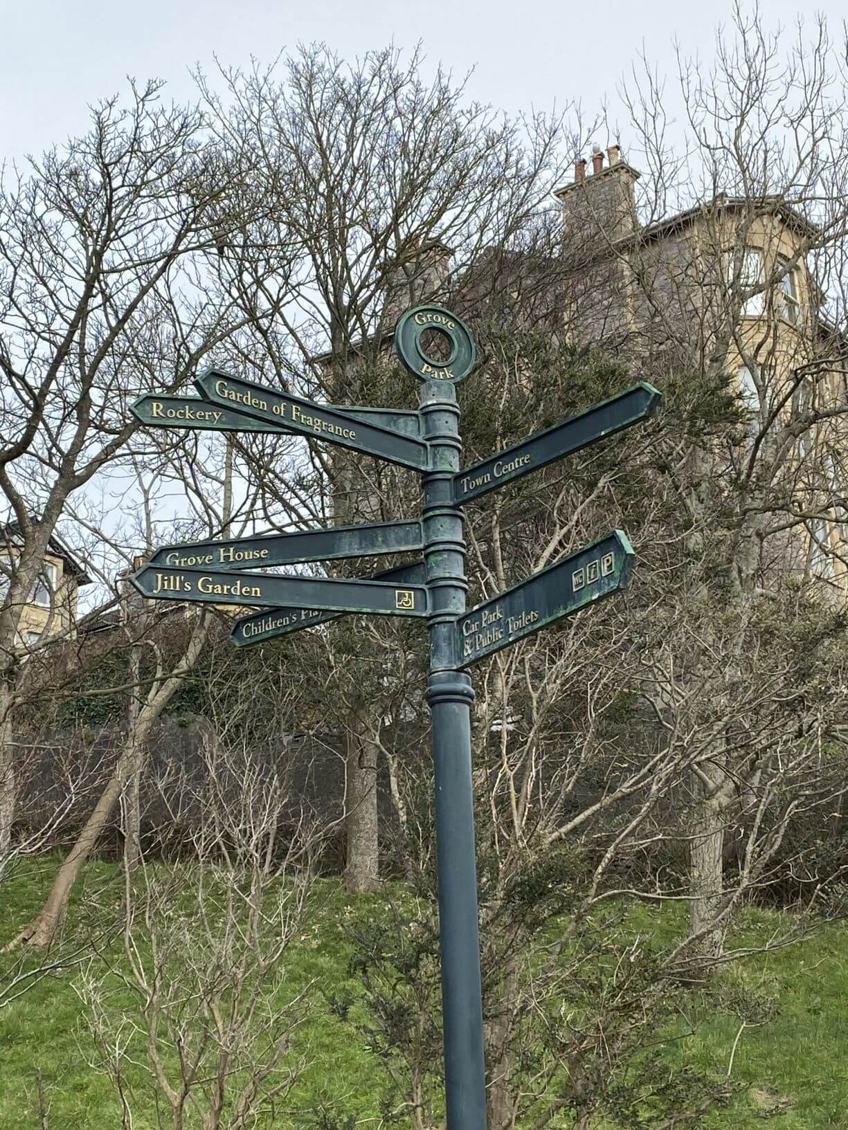 A multi-directional signpost in a park with directions to the Rockery, Town Center, Grove House, Jill's Garden, and other sites. - Home Instead