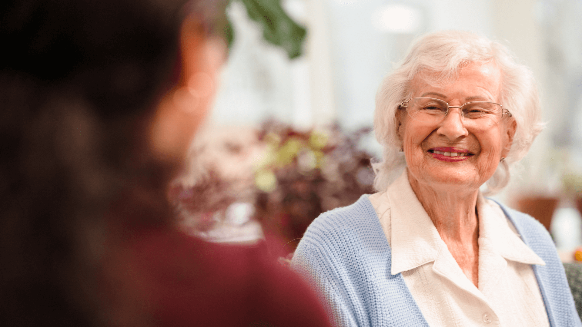 Elderly woman with white hair and glasses smiling warmly at another person in an indoor setting. - Home Instead