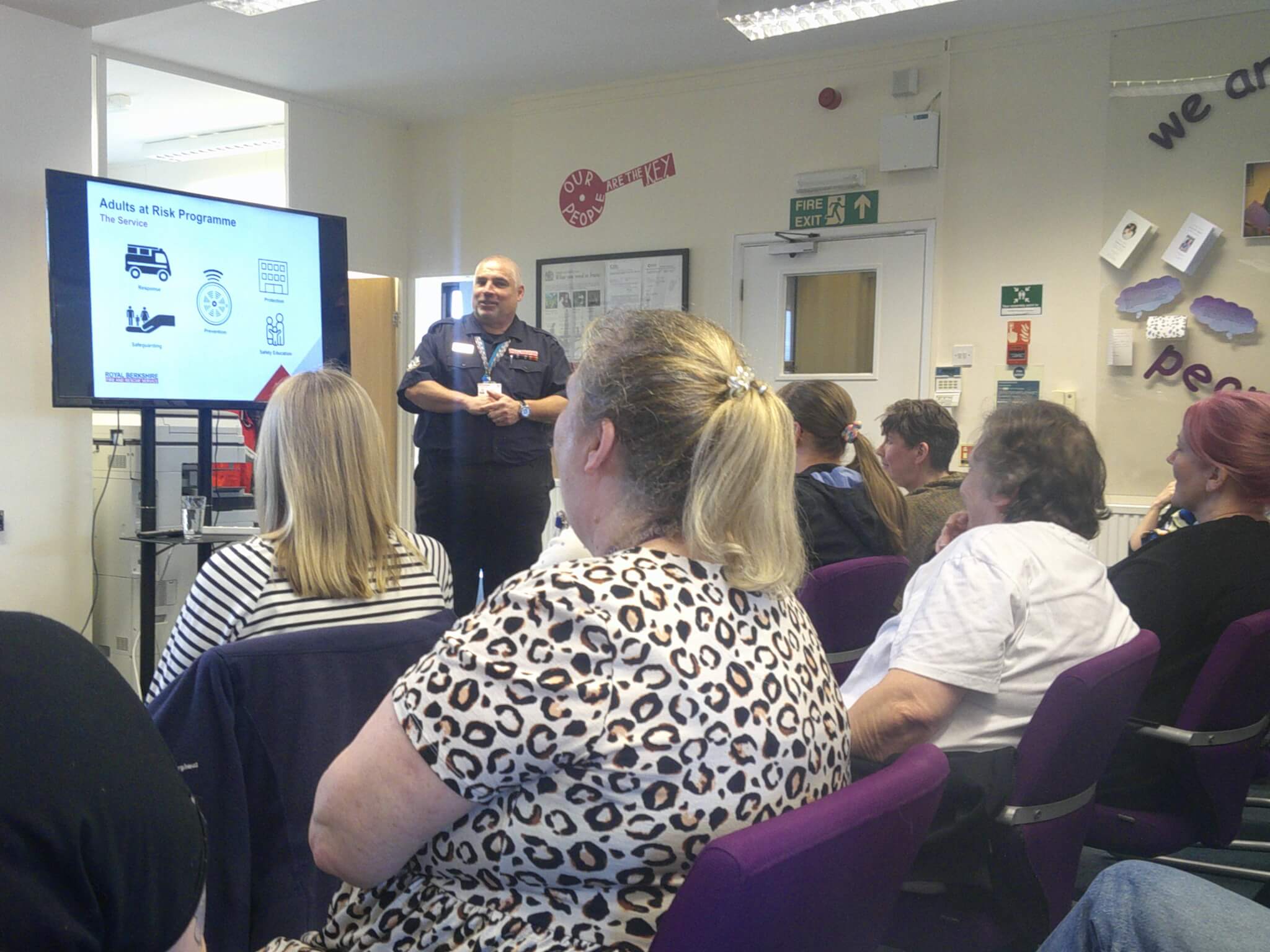 A presenter talks to an attentive audience seated in a room; a slide on "Adults at Risk Programme" is displayed on a screen. - Home Instead