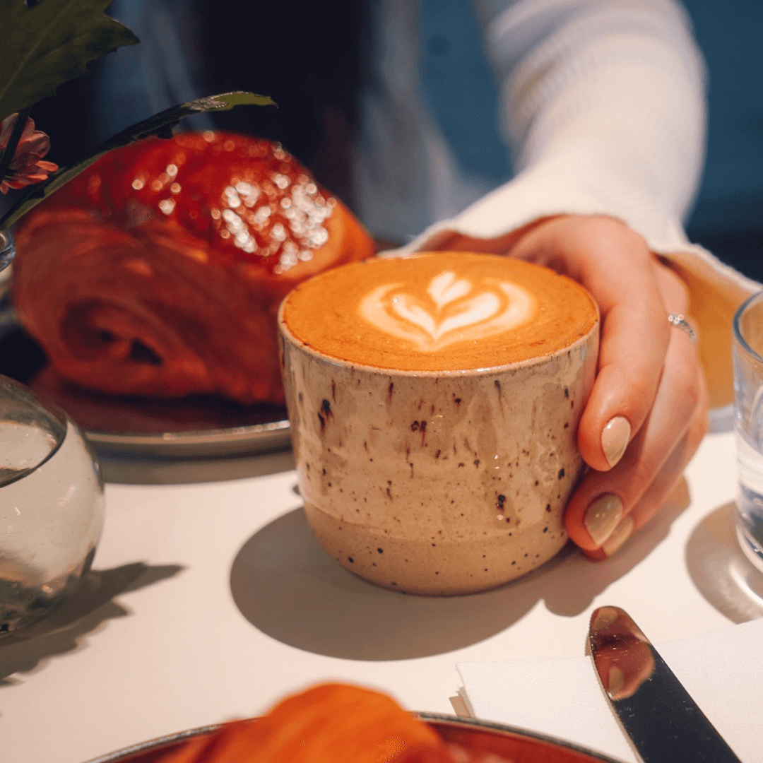 A hand holding a latte with latte art, next to a croissant on a table with a small vase. - Home Instead