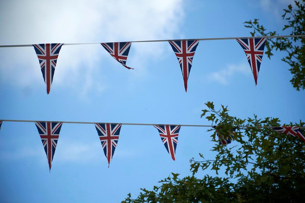 Two strings of Union Jack bunting flags against a blue sky with a few clouds and some tree branches on the right. - Home Instead