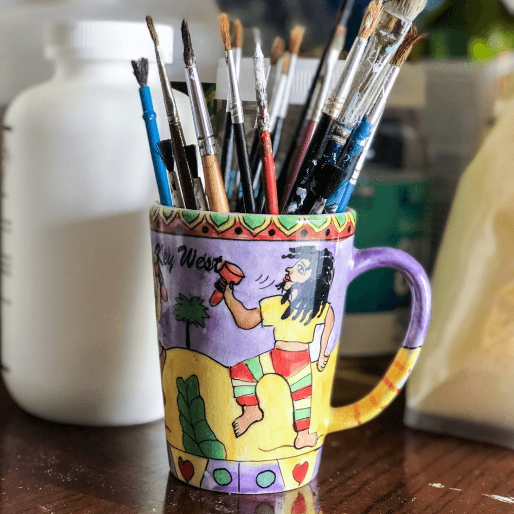 A colorful mug decorated with a painting holds various paintbrushes, placed on a table surrounded by art supplies. - Home Instead