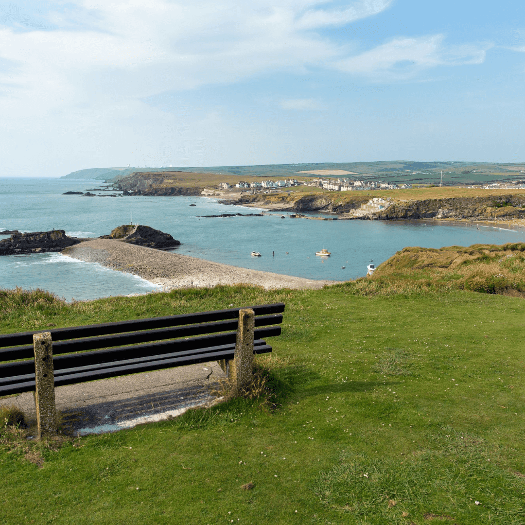 A bench on a grassy hill overlooks a coastal landscape with cliffs, a beach, and a small village in the distance. - Home Instead