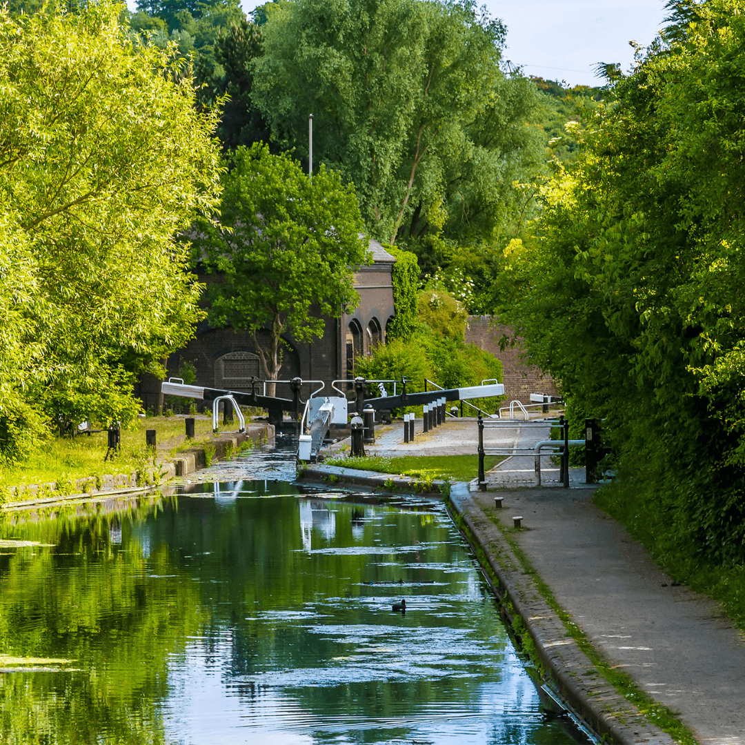 A serene canal lined with trees and greenery, featuring a lock gate and brick buildings in the background. - Home Instead