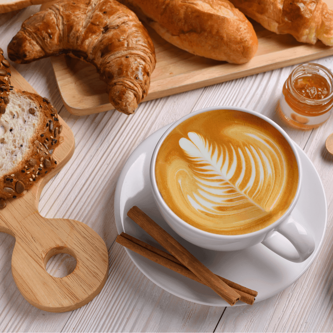 A latte with art in a white cup, cinnamon sticks, croissants, sliced bread, and a small jar of marmalade on a table. - Home Instead