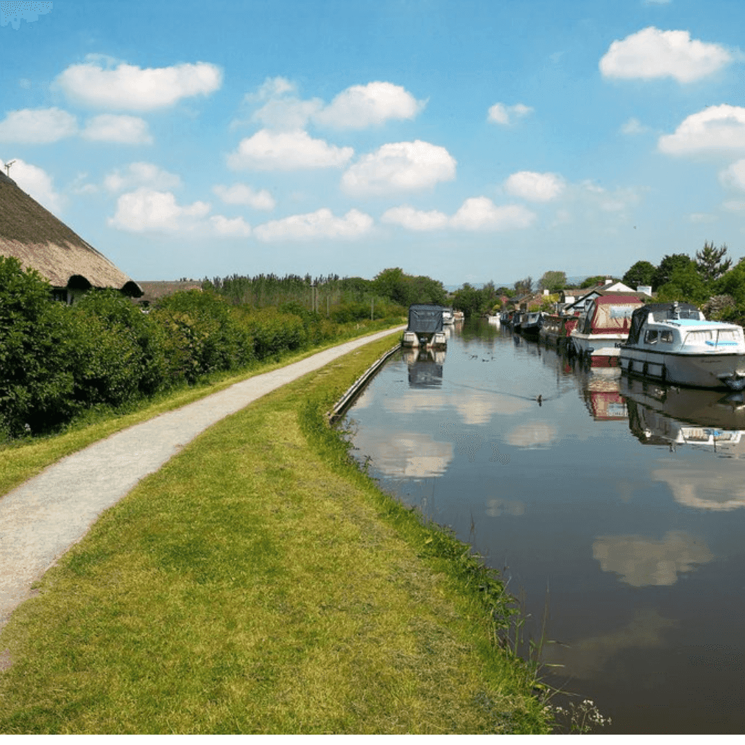 A peaceful canal scene with boats moored on the right and a pathway with greenery on the left under a blue sky with clouds. - Home Instead