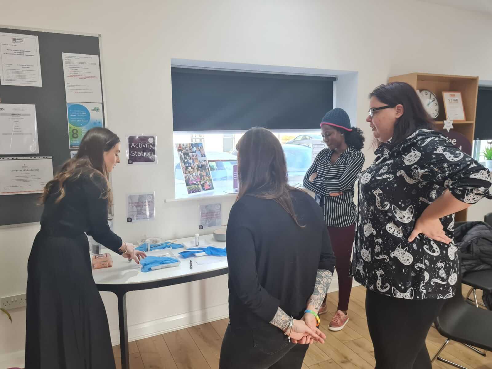Four women standing around a table with medical supplies, engaging in a hands-on activity in a well-lit room. - Home Instead