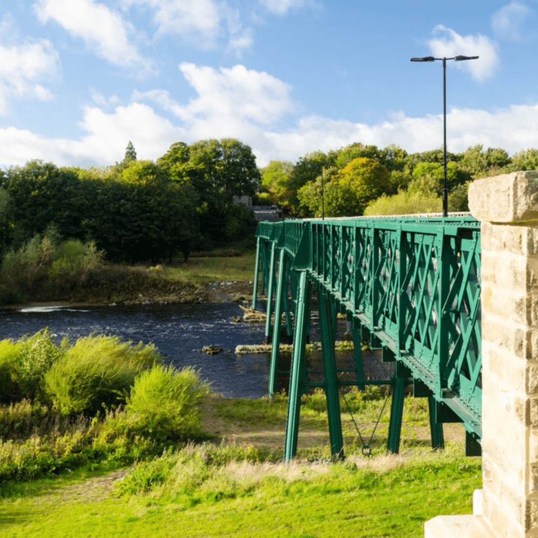 A green metal bridge stretches over a river, surrounded by lush greenery and under a partly cloudy blue sky. - Home Instead