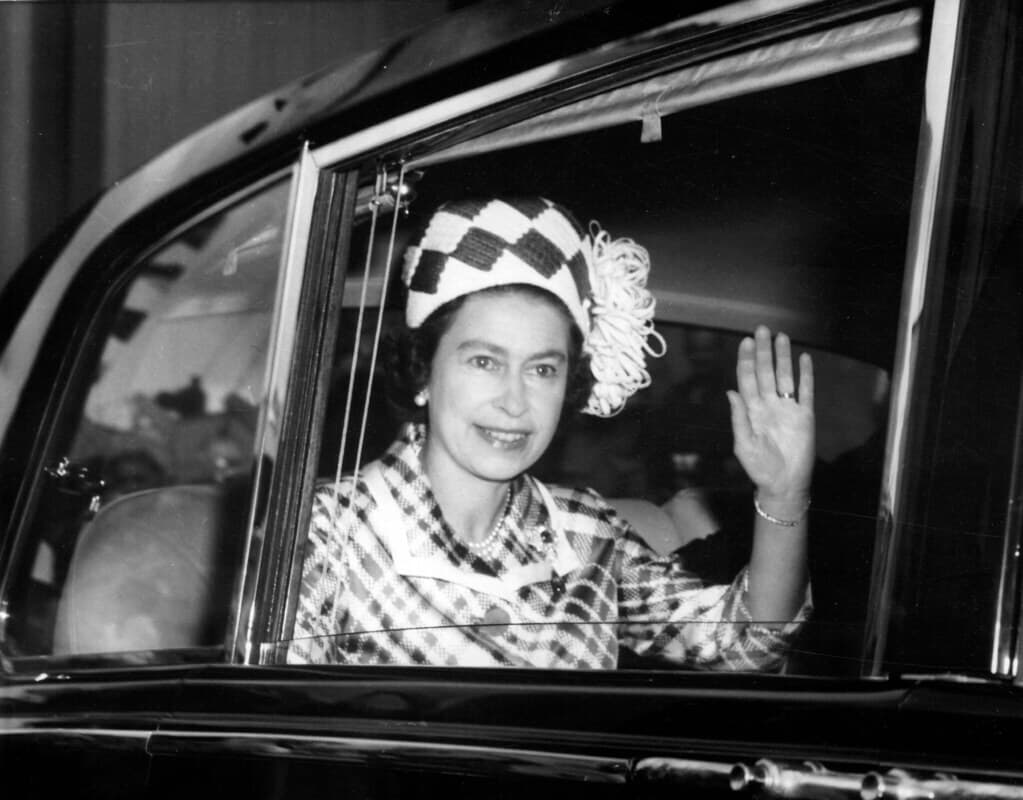 A woman in a plaid outfit and hat waves from a car window with a gentle smile on her face. - Home Instead