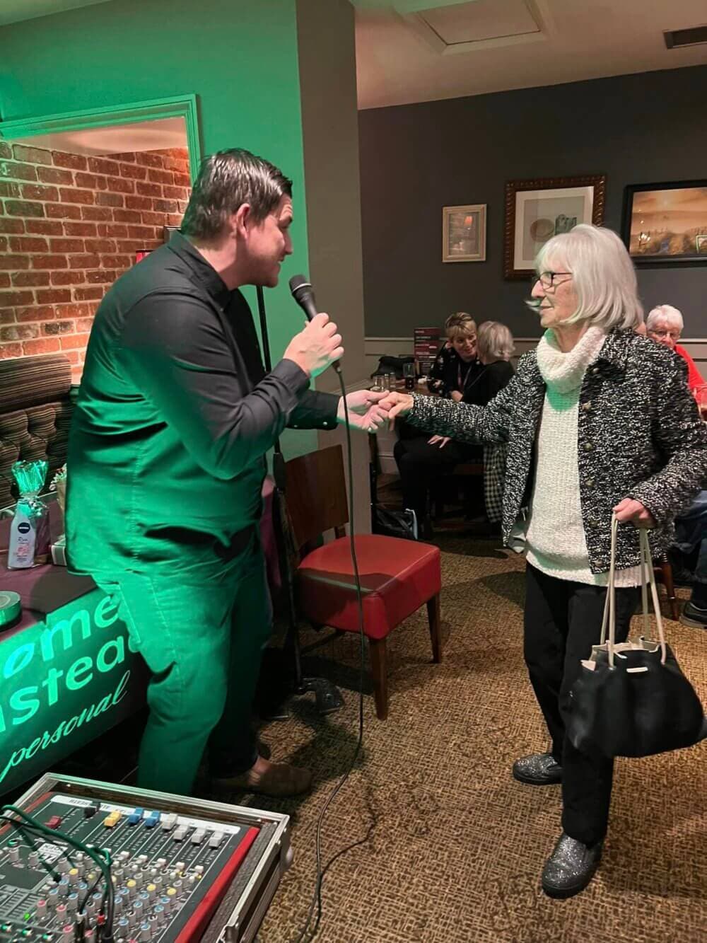 A man with a microphone and an elderly woman shaking hands in a warmly lit indoor setting. - Home Instead