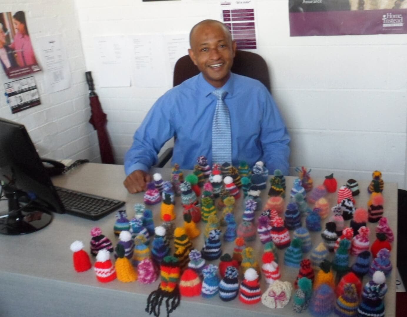 Smiling man in an office sits behind a desk with a large collection of colorful, knitted mini hats displayed. - Home Instead