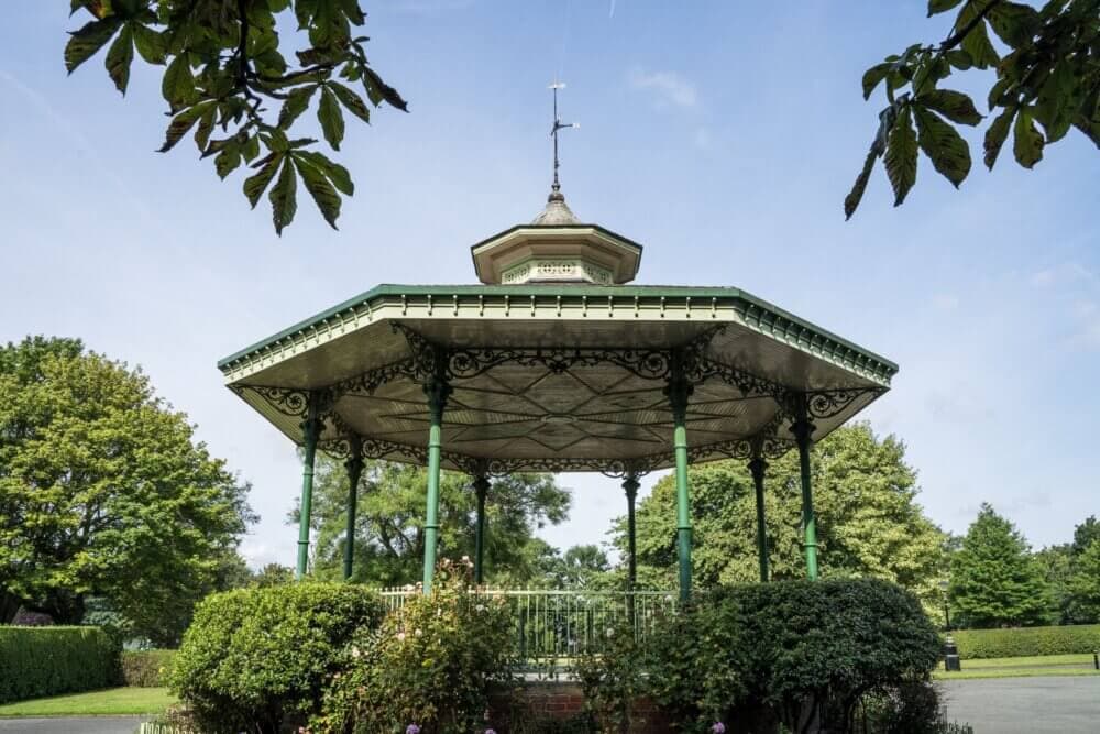 A green, octagonal bandstand in a park, surrounded by lush trees and bushes, under a clear blue sky. - Home Instead