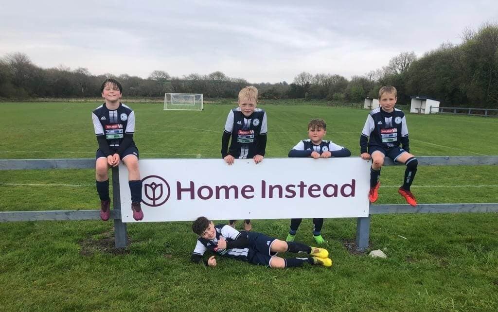 Five young soccer players in uniform pose around a "Home Instead" sign on a grassy field. - Home Instead