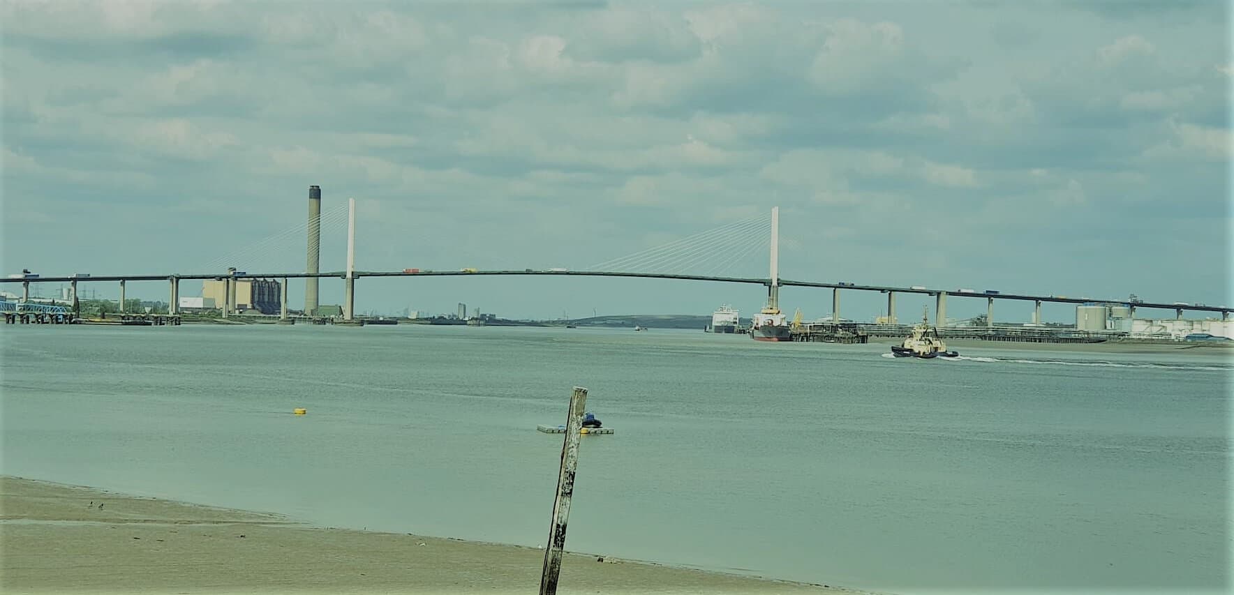 Wide view of a large suspension bridge over a calm river with boats and cloudy sky in the background. - Home Instead