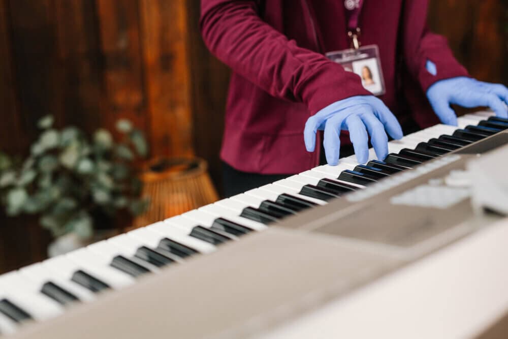 Person in gloves playing a keyboard piano indoors with a plant and a wooden wall in the background. - Home Instead