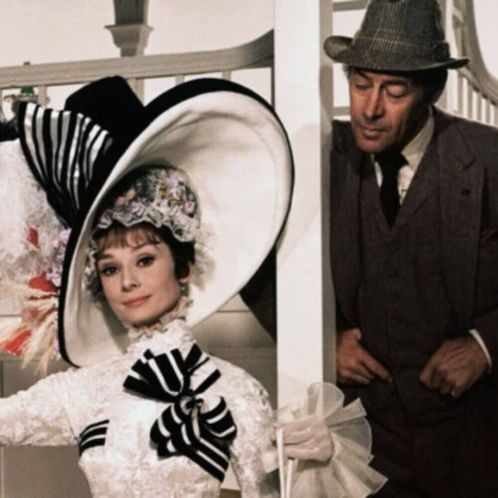 A woman in an elegant striped hat and dress is observed by a man in a suit and hat in a classic film scene. - Home Instead