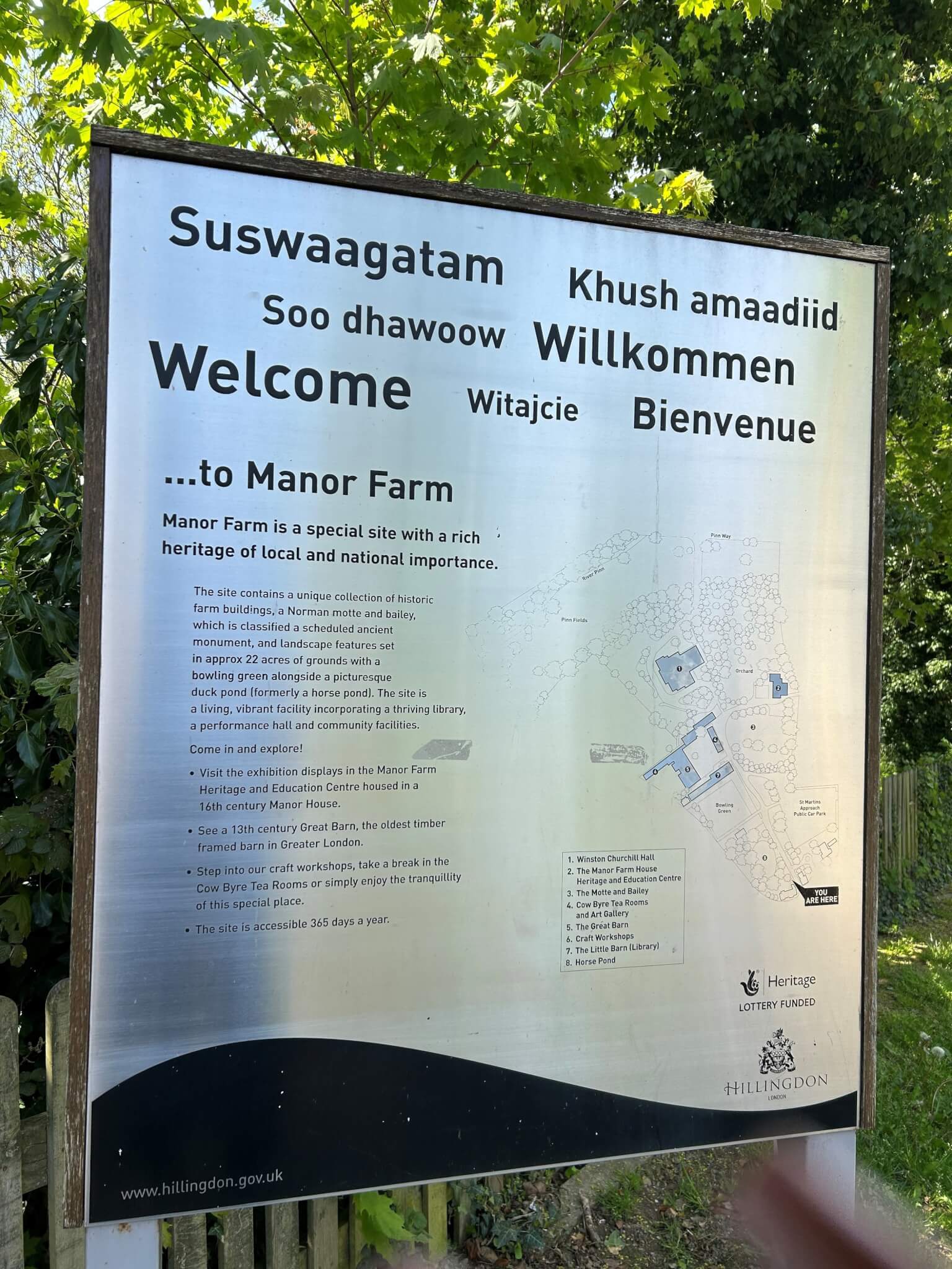 A multilingual welcome sign for Manor Farm, with a site map and brief description, surrounded by green foliage. - Home Instead
