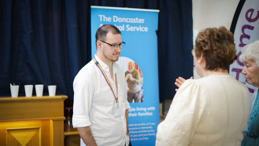 A man engages in conversation with two women near a banner for "The Doncaster Admiral Service. - Home Instead