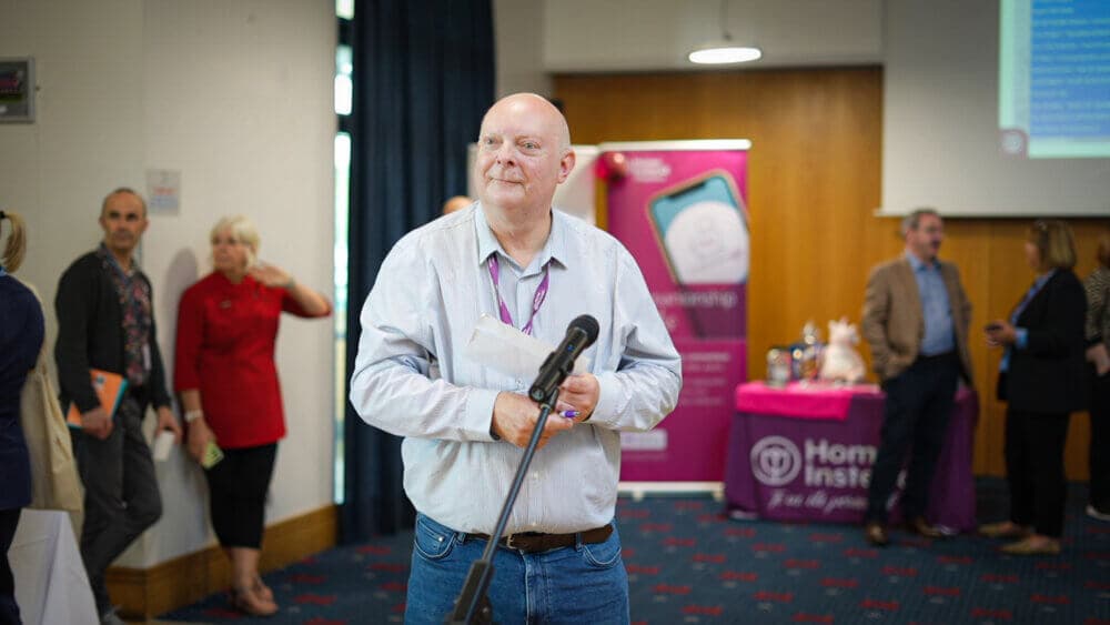 A man in a light blue shirt speaks into a microphone at an event with people and banners in the background. - Home Instead