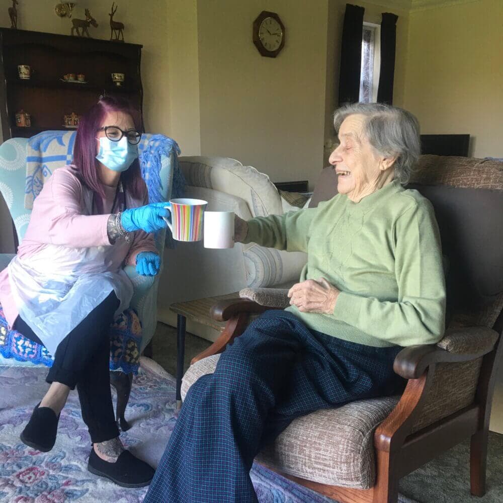 A caregiver wearing a mask and gloves shares a cheerful moment, clinking mugs with an elderly woman in a cozy living room. - Home Instead