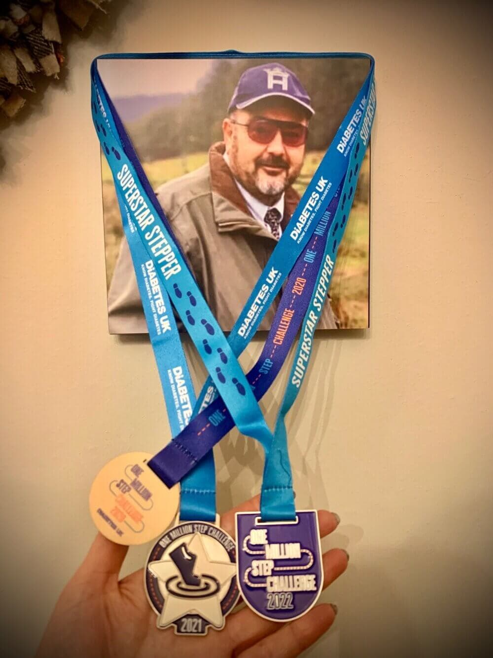 A hand holding three medals with blue lanyards in front of a framed photo of a man wearing sunglasses and a hat. - Home Instead