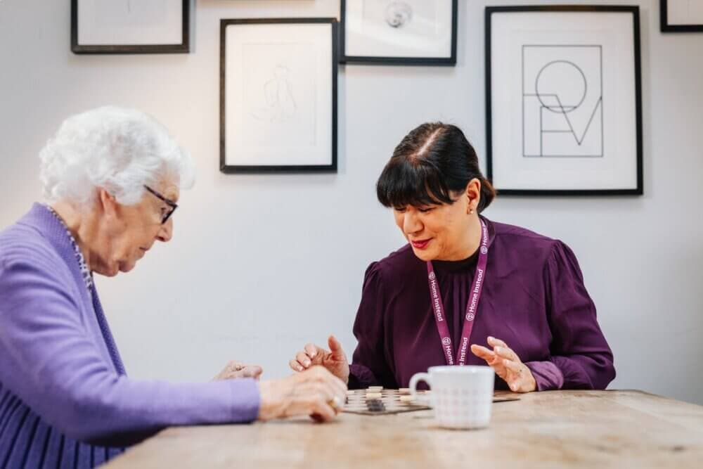 Two women sitting at a table, engaged in a checkerboard game, with framed artwork on the wall behind them. - Home Instead Poole