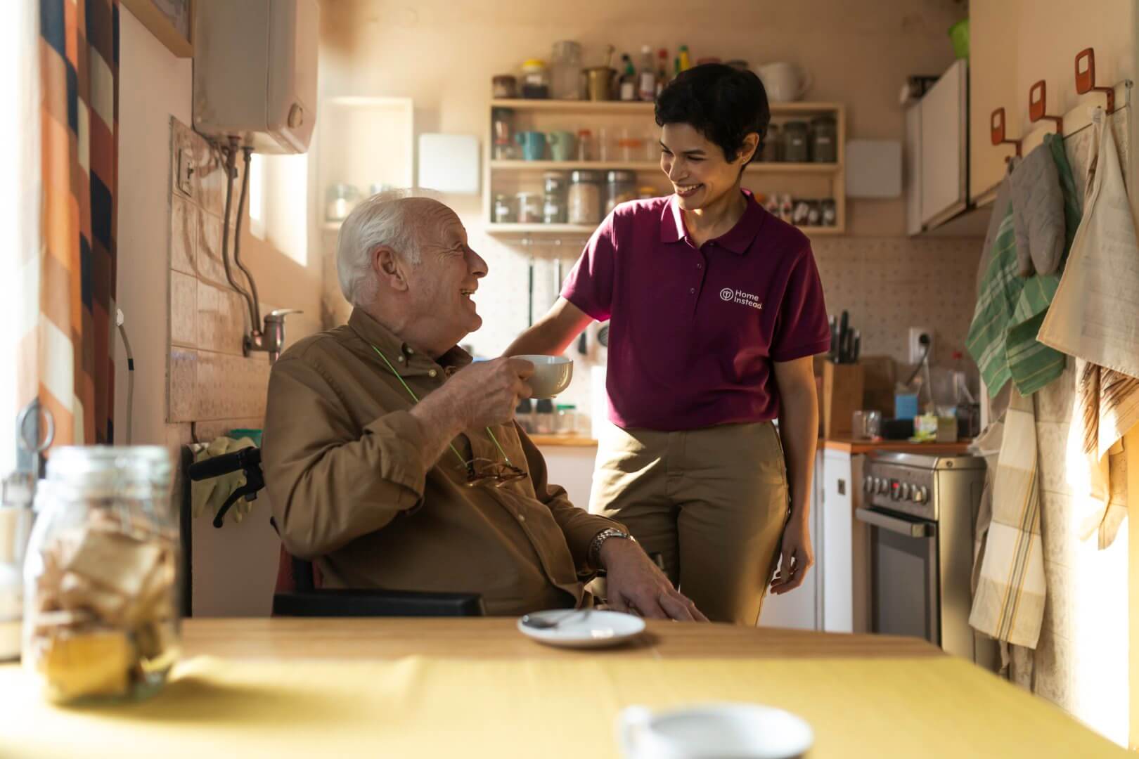 A Care Professional in a purple shirt smiles at an elderly man holding a cup at a kitchen table. They appear to be conversing happily. - Home Instead Bournemouth & Christchurch