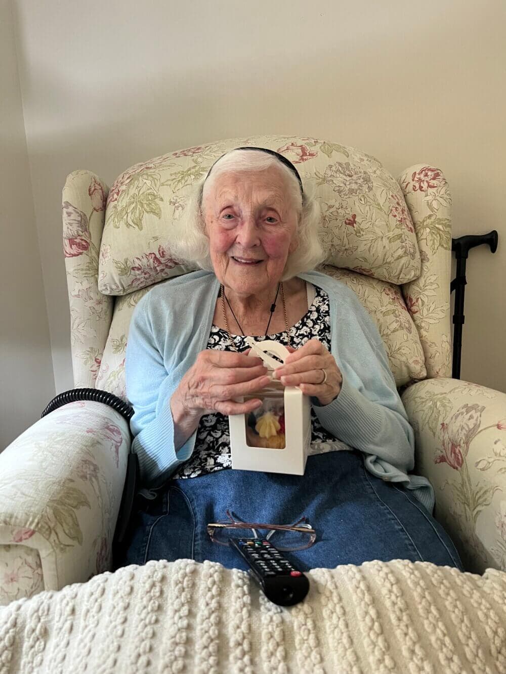 An elderly woman with white hair, seated in a floral armchair, smiling and holding a small box. - Home Instead