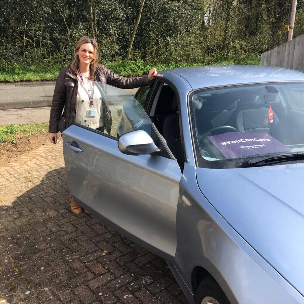 Woman standing next to an open car door, smiling, with a badge around her neck and #YouDeserveCare sign on the windshield. - Home Instead