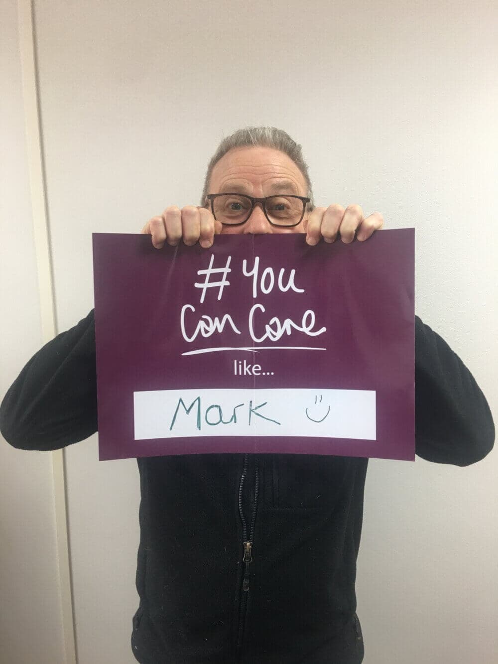 Person holding a purple sign that reads "You can care like Mark" with a smiley face, partially covering their face. - Home Instead