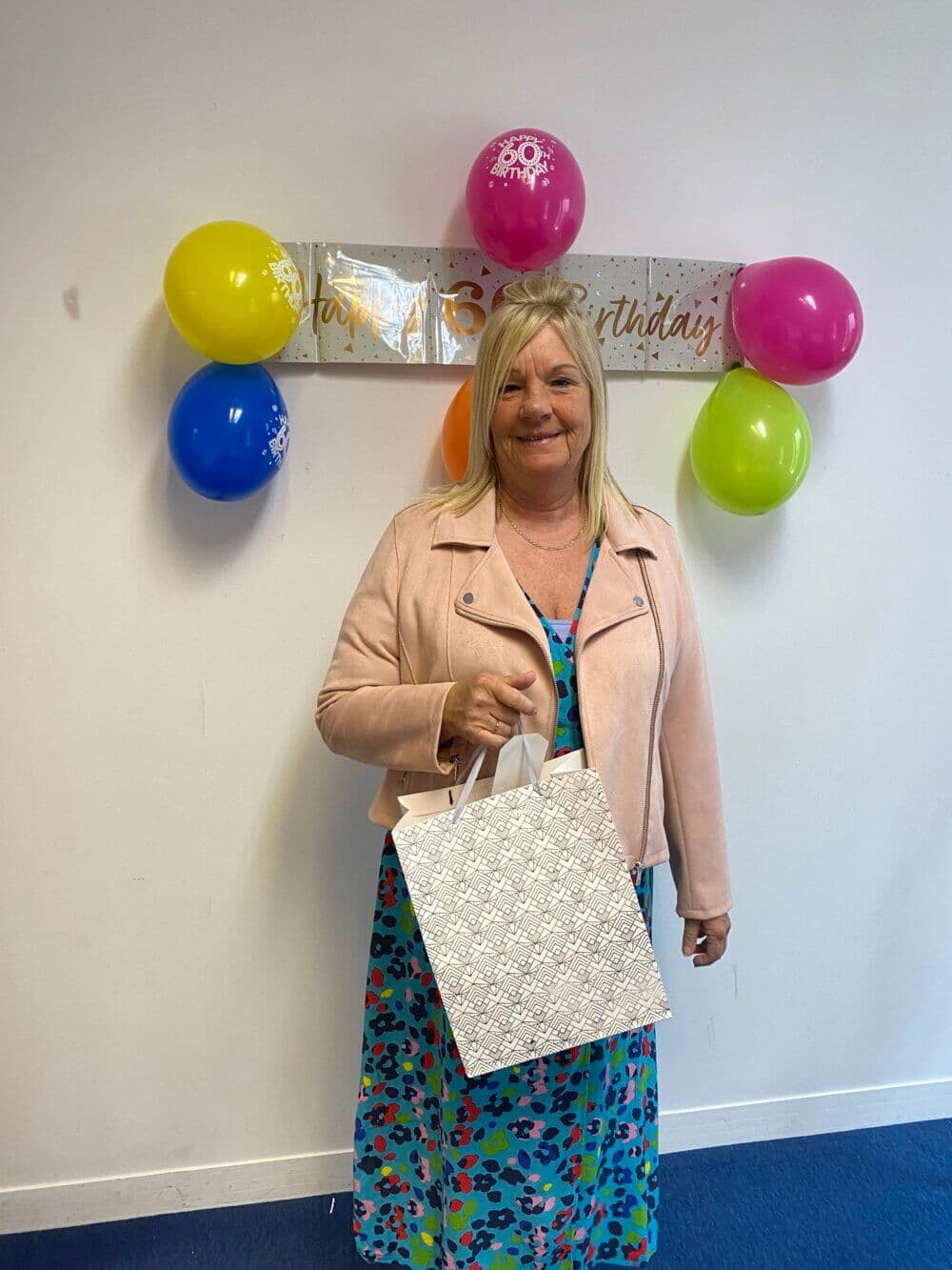 A woman holding a gift bag stands in front of a "Happy Birthday" banner and colorful balloons. - Home Instead