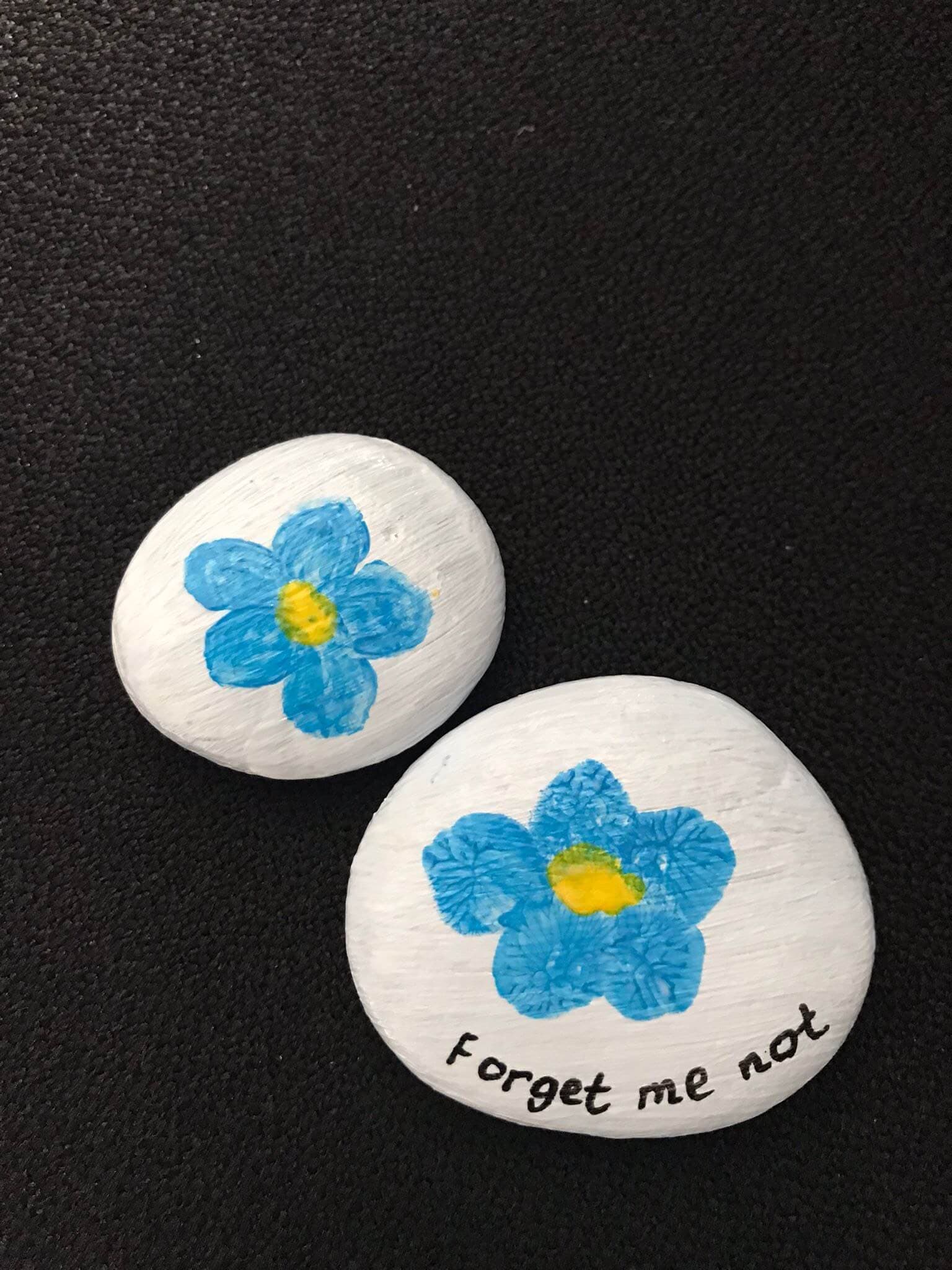 Two white stones with blue flowers, one with "forget me not" text, placed on a black surface. - Home Instead