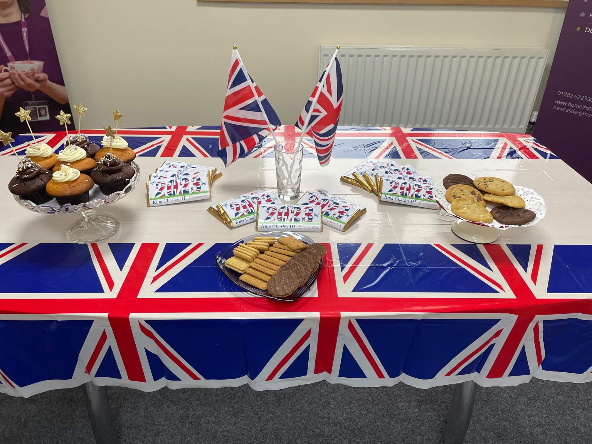 A table decorated with Union Jack flags and covers, featuring cakes, biscuits, and snacks. - Home Instead