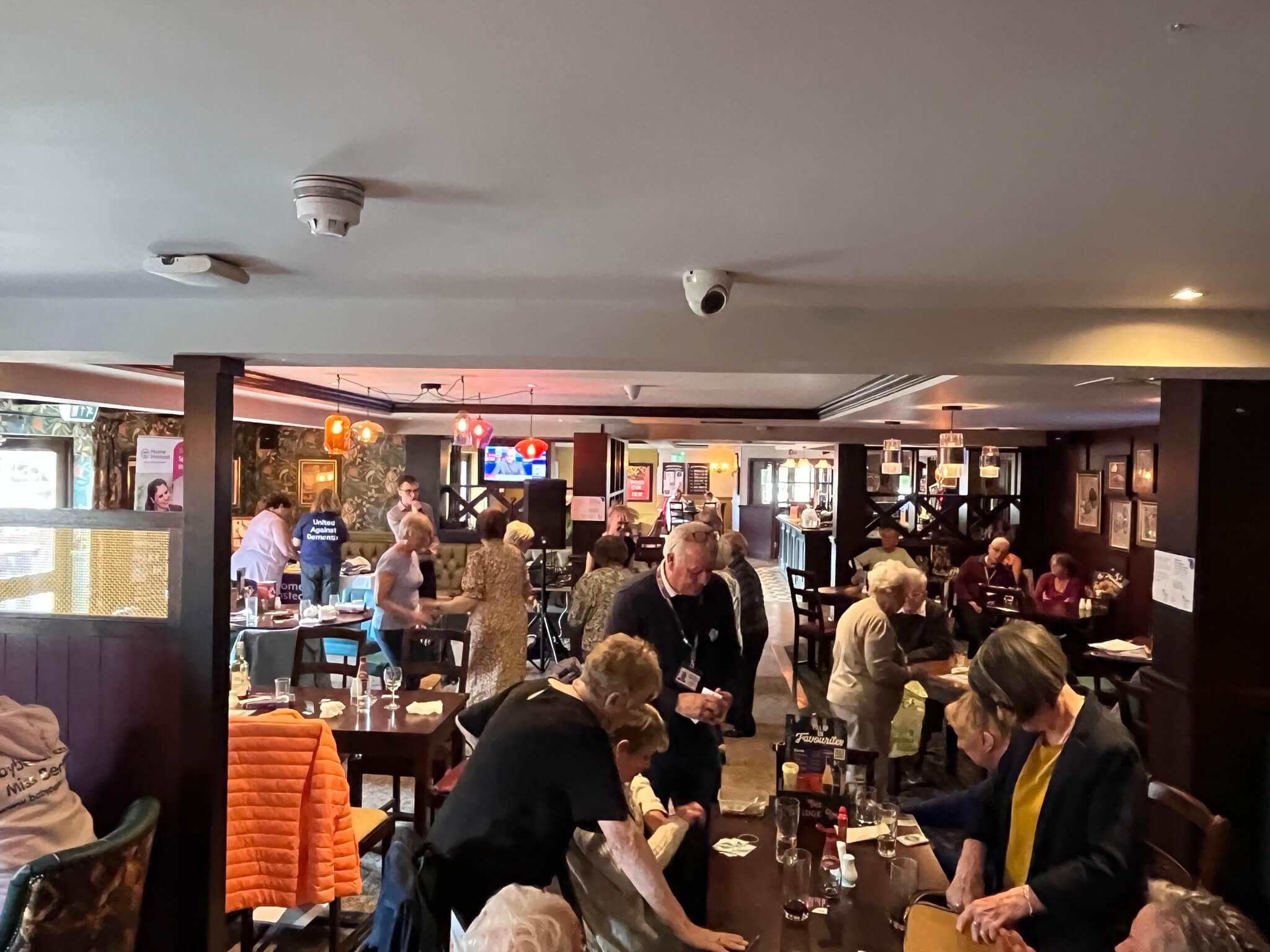 A group of people socializing in a busy restaurant or pub with tables and various activities taking place. - Home Instead