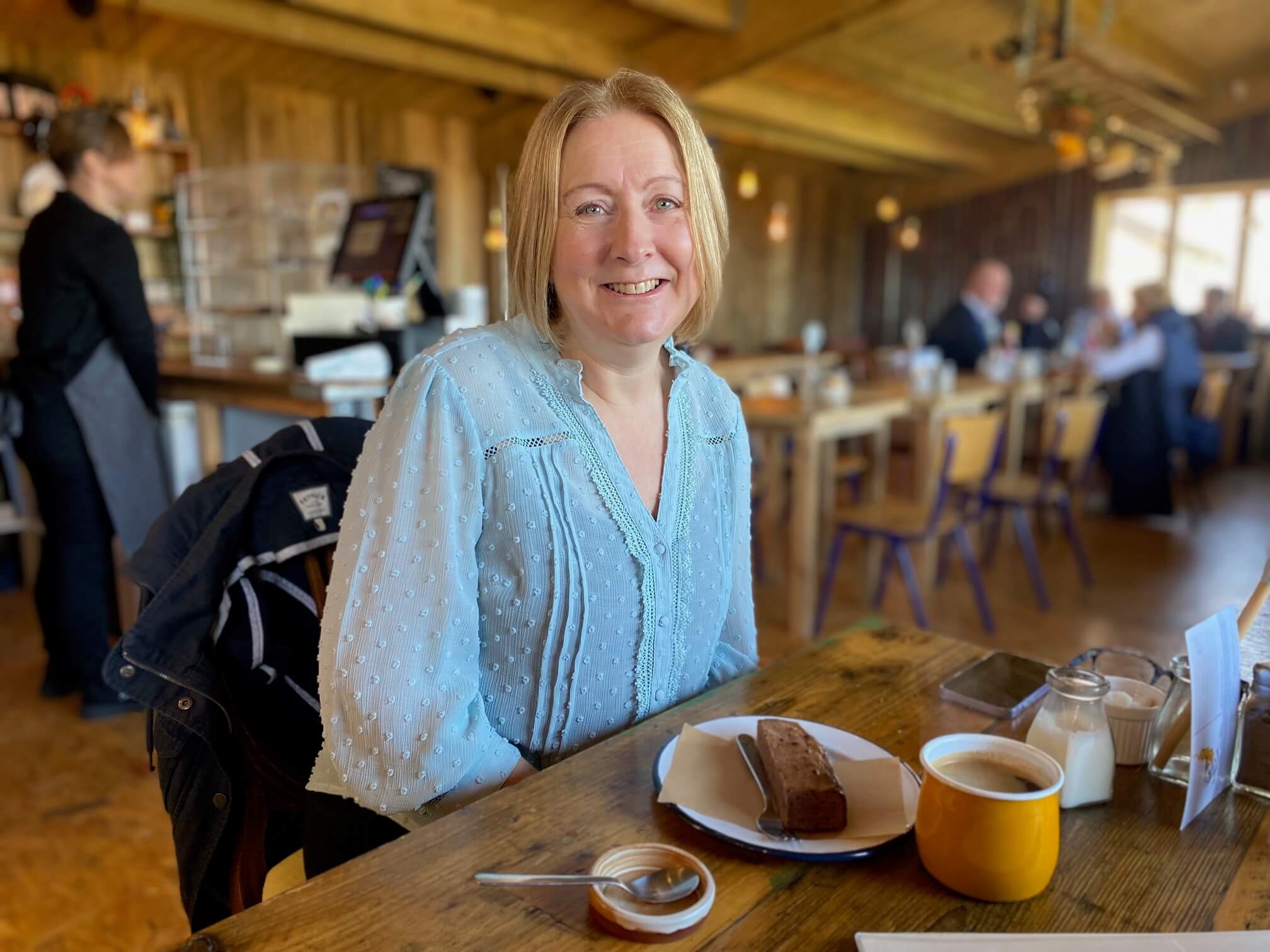 A person smiling while seated at a wooden table with a slice of cake and a cup of coffee in a cozy café. - Home Instead