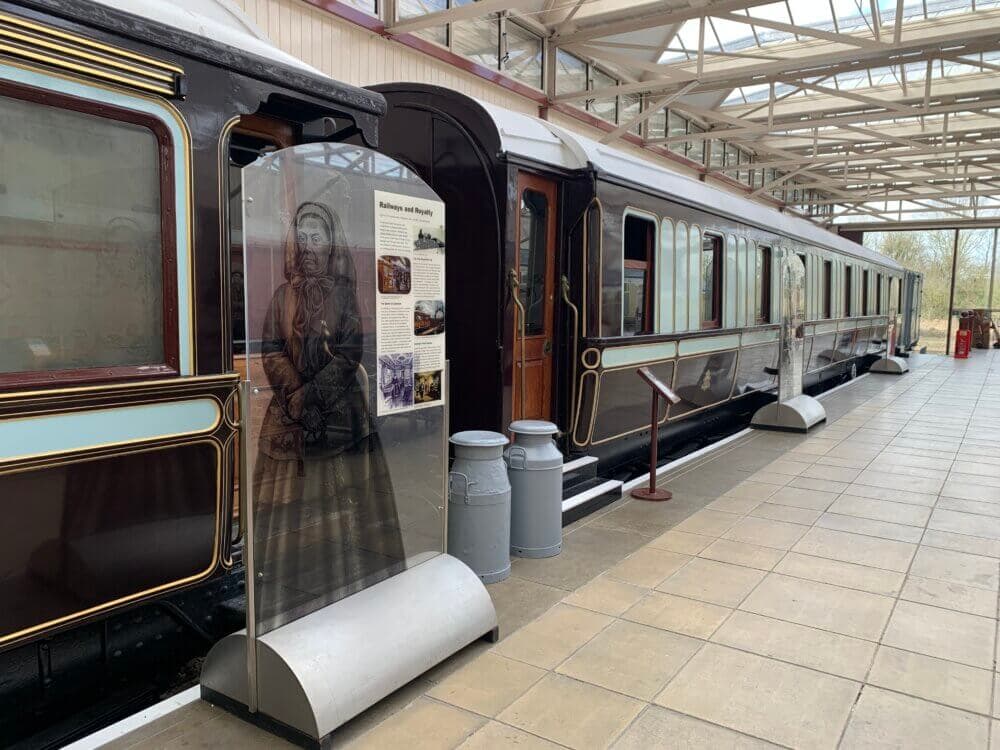 A historic train car displayed in a sheltered outdoor area, with an informational sign and milk churns nearby. - Home Instead