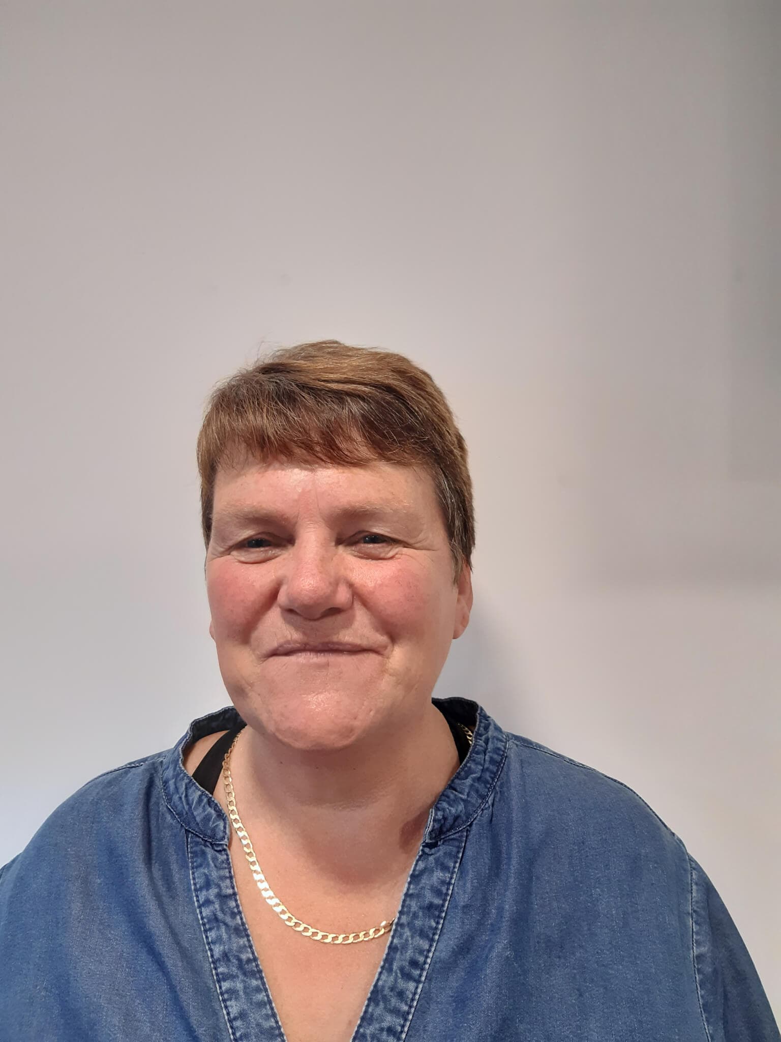 Smiling person with short hair, wearing a denim top and a necklace, standing against a plain light-colored background. - Home Instead